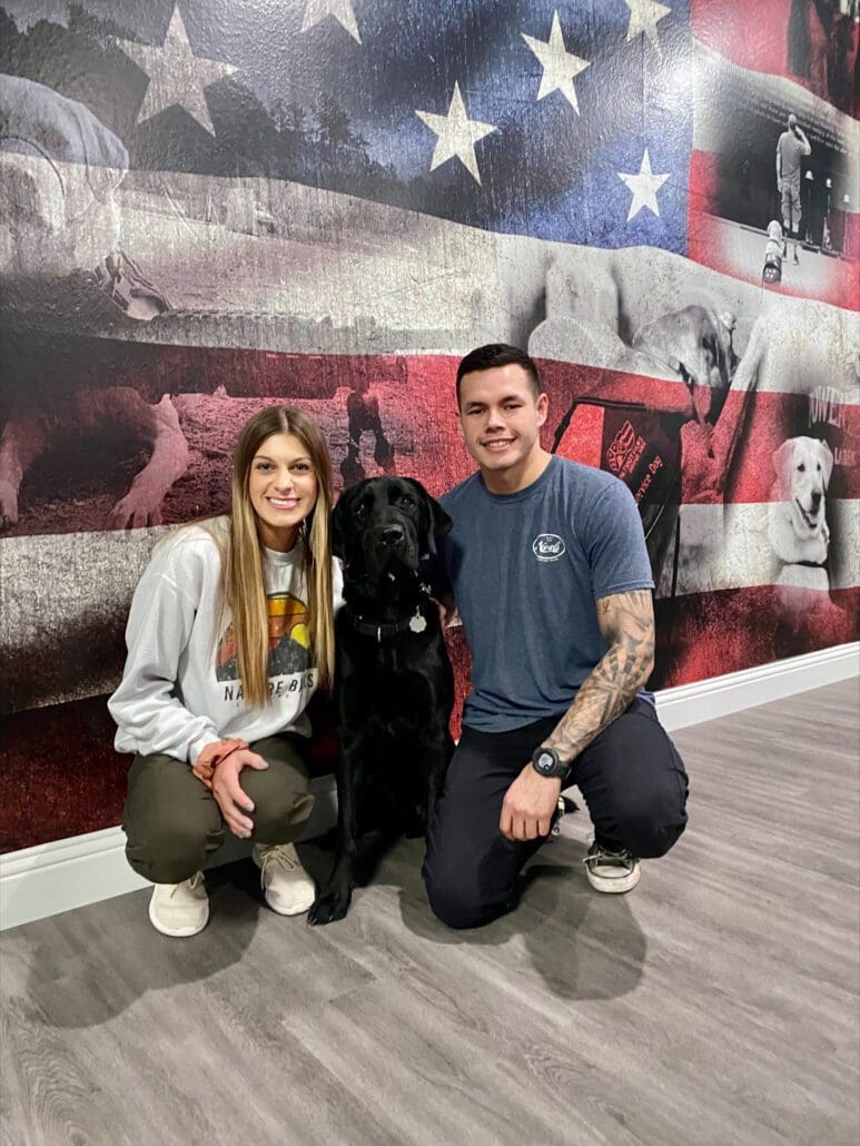 A man and woman kneeling next to a black dog.